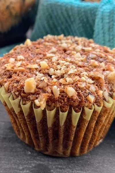 Close up of a banana nut muffin topped with walnuts and a blue cloth lined basket of muffins in the background.