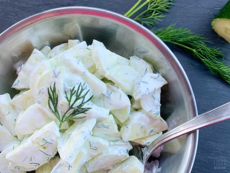 A metal bowl of creamy cucumber salad with fresh dill sitting on a slate board. A few springs of fresh dill and a half of a cucumber is resting nearby.